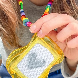 May include: A yellow drawing toy with a white screen and a gray heart design is held up by a rainbow beaded necklace. The necklace features a variety of colorful beads. The toy has a clear yellow frame.