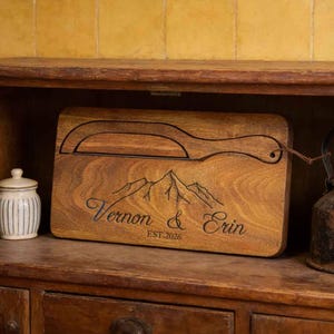 May include: A wooden cutting board with a cheese knife design and the names "Vernon & Erin" engraved, along with the year "EST. 2026". The board is displayed inside a wooden cabinet with a vintage kettle and two small jars.