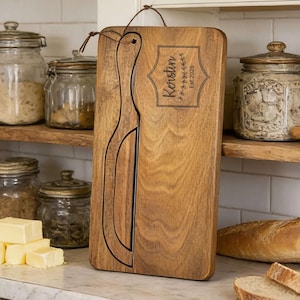 Puede incluir: Una tabla de cortar de madera con un diseño único en forma de botella se muestra en una cocina. La tabla presenta el nombre "Kerstin" y el año "Est. 2020". También se ven tarros de ingredientes y una barra de pan.