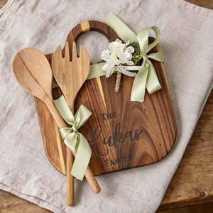 Op de afbeelding: Een houten serveerplank met handvat, een houten lepel en een houten vork samengebonden met een lichtgroen lint. De plank is gegraveerd met de tekst The Lukas Family en versierd met een klein boeketje van witte kunstbloemen.
