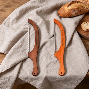 May include: Two wooden bread knives with serrated blades. One knife is a dark brown color, and the other is orange. The text "Best Time, Bread Time" is engraved on the wooden handles. A loaf of bread and a linen cloth are in the background.