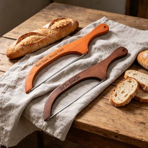 May include: Two wooden bread slicers with curved blades rest on a linen cloth. One is light brown, the other dark brown. The text "BEST TIME, BREAD TIME" is engraved on the light brown slicer. Sliced bread and a loaf of bread are also visible.
