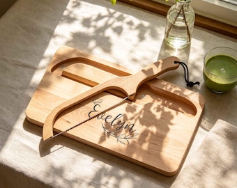Könnte beinhalten: Ein rechteckiges Käsebrett aus hellem Holz mit einem passenden Käsehobel aus Holz, der mit einem dünnen Metalldraht ausgestattet ist. Das Brett ist mit dem Namen Evelyn, einem kleinen Kochmützen-Symbol und einem Blattzweig-Motiv graviert.