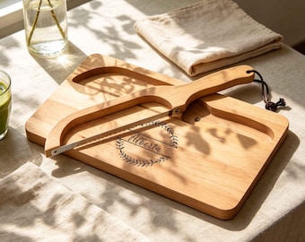 Könnte beinhalten: Ein rechteckiges Schneidebrett aus hellem Holz mit einer eingefrästen Rille und einem passenden Käsehobel aus Holz mit Metalldraht. Das Brett ist mit einem kreisförmigen Blattkranz-Motiv graviert, das den Namen Alberta in Schreibschrift umschließt.
