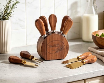 Könnte beinhalten: Ein Käsemesserset mit vier goldfarbenen Metallklingen und dunkelbraunen Holzgriffen. Das Set enthält einen runden Holzblock mit der Gravur Chantelle Est. 2020, der drei Messer hält, während eine Gabel und zwei weitere Messer separat auf der Fläche liegen.