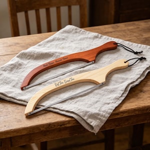 May include: Two wooden bread saws with curved handles and serrated blades rest on a linen cloth. One is a rich reddish-brown, the other a light natural wood. The text "Best Time, Bread Time" is etched on each handle.
