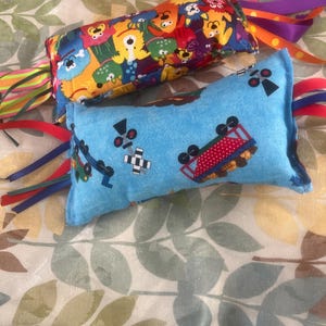 May include: Two rectangular sensory toys. One is blue with train and crossing graphics. The other is patterned with colorful cartoon characters. Both have colorful ribbons attached to the ends.