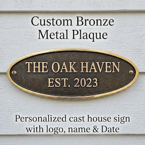 May include: An oval bronze metal plaque with a dark textured center and a polished gold-toned rim. The plaque features the raised text THE OAK HAVEN and EST. 2023 in a gold-toned finish. It is secured with two visible screws on the left and right sides.