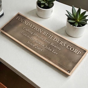Puede incluir: Placa rectangular de bronce con el texto "FOUNDATION BUILDERS CORP" y "Crafting Dreams Into Reality". También incluye "Established by Sarah & John Lee Since 2005". La placa está enmarcada con un borde de bronce más claro.