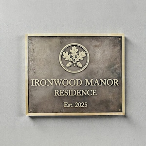 Puede incluir: Un letrero metálico rectangular de color bronce con un borde dorado. El letrero presenta el texto "IRONWOOD MANOR RESIDENCE" y "Est. 2025" en letras doradas. Un emblema circular con hojas de roble y bellotas en la parte superior.