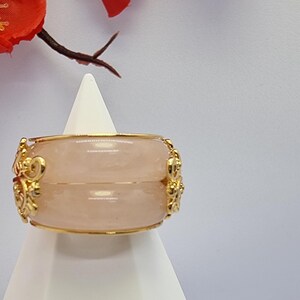 May include: A gold-toned bracelet with a light pink stone is displayed on a white stand. The bracelet features ornate gold detailing and is set against a backdrop of red flowers and a white background. The jewelry is a decorative accessory.