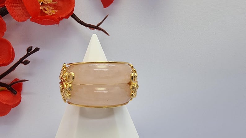 May include: A gold-toned bracelet with a light pink stone is displayed on a white stand. The bracelet features ornate gold detailing and is set against a backdrop of red flowers and a white background. The jewelry is a decorative accessory.