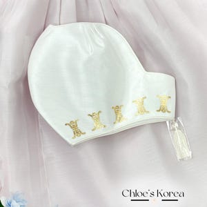 Peut inclure: Chapeau en satin blanc avec un motif brodé doré et un pompon blanc. Le chapeau est sur un fond rose.