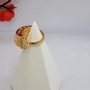 May include: A gold-colored ring with an ornate design, featuring intricate details and embellishments. The ring is displayed on a white geometric stand, with red floral accents in the background. The ring's design includes a detailed sculpted element.
