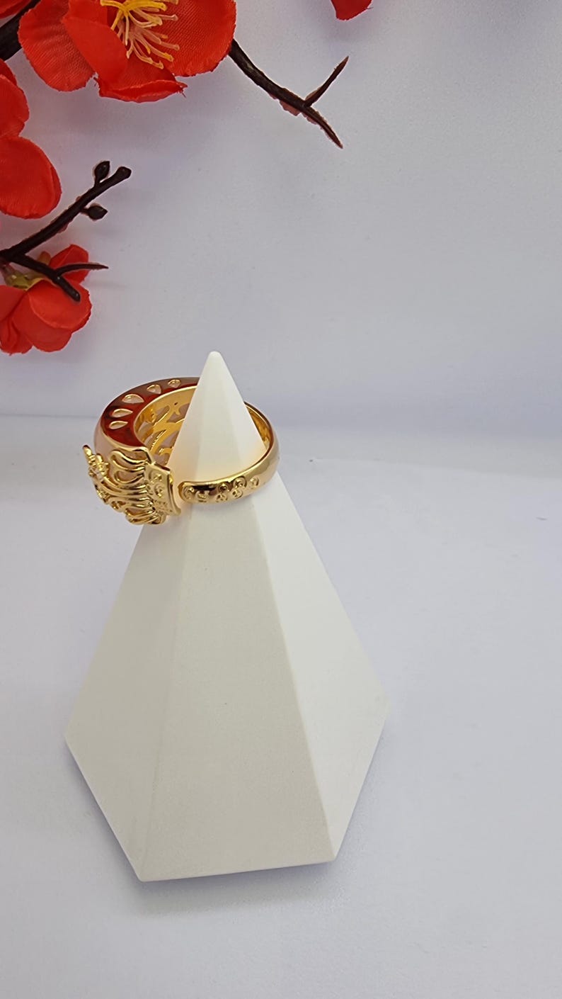 May include: A gold-colored ring with an ornate design, featuring intricate details and embellishments. The ring is displayed on a white geometric stand, with red floral accents in the background. The ring's design includes a detailed sculpted element.