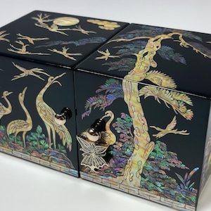 Puede incluir: Caja lacada en negro con intrincados incrustaciones de nácar. La caja presenta un diseño de paisaje con grullas, árboles y pájaros. La caja tiene una tapa con bisagras y un pomo decorativo. La caja mide aproximadamente 15 cm de ancho, 15 cm de profundidad y 10 cm de alto.