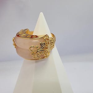 May include: A wide, rose-colored ring with gold filigree detailing. The ring is displayed on a white, geometric stand. The gold filigree wraps around the pink stone, creating an ornate design. The ring is a statement piece.