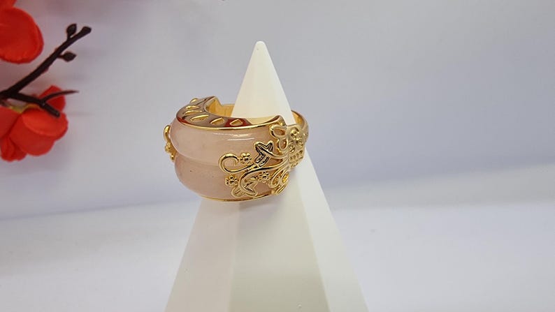 May include: A wide, rose-colored ring with gold filigree detailing. The ring is displayed on a white, geometric stand. The gold filigree wraps around the pink stone, creating an ornate design. The ring is a statement piece.