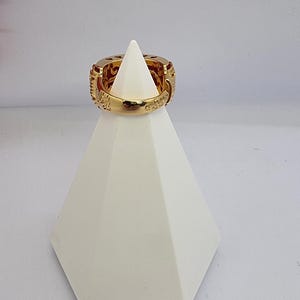 May include: A gold-colored ring with intricate carvings and a wide band is displayed on a white geometric stand. The ring's design features detailed patterns and textures, showcasing a luxurious aesthetic. The ring is a statement piece.