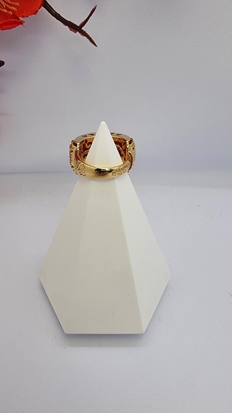 May include: A gold-colored ring with intricate carvings and a wide band is displayed on a white geometric stand. The ring's design features detailed patterns and textures, showcasing a luxurious aesthetic. The ring is a statement piece.
