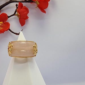 May include: A wide, light pink ring with gold filigree detailing. The ring is displayed on a white stand, with red flowers and branches in the background. The ring's design is symmetrical, with ornate gold accents.