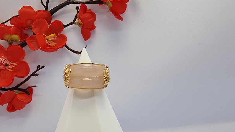 May include: A wide, light pink ring with gold filigree detailing. The ring is displayed on a white stand, with red flowers and branches in the background. The ring's design is symmetrical, with ornate gold accents.