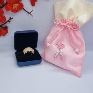 May include: A gold ring with a light pink stone is displayed in a blue ring box. A pink satin gift bag with a cream-colored top is next to the ring box. Red flowers and branches are in the background.