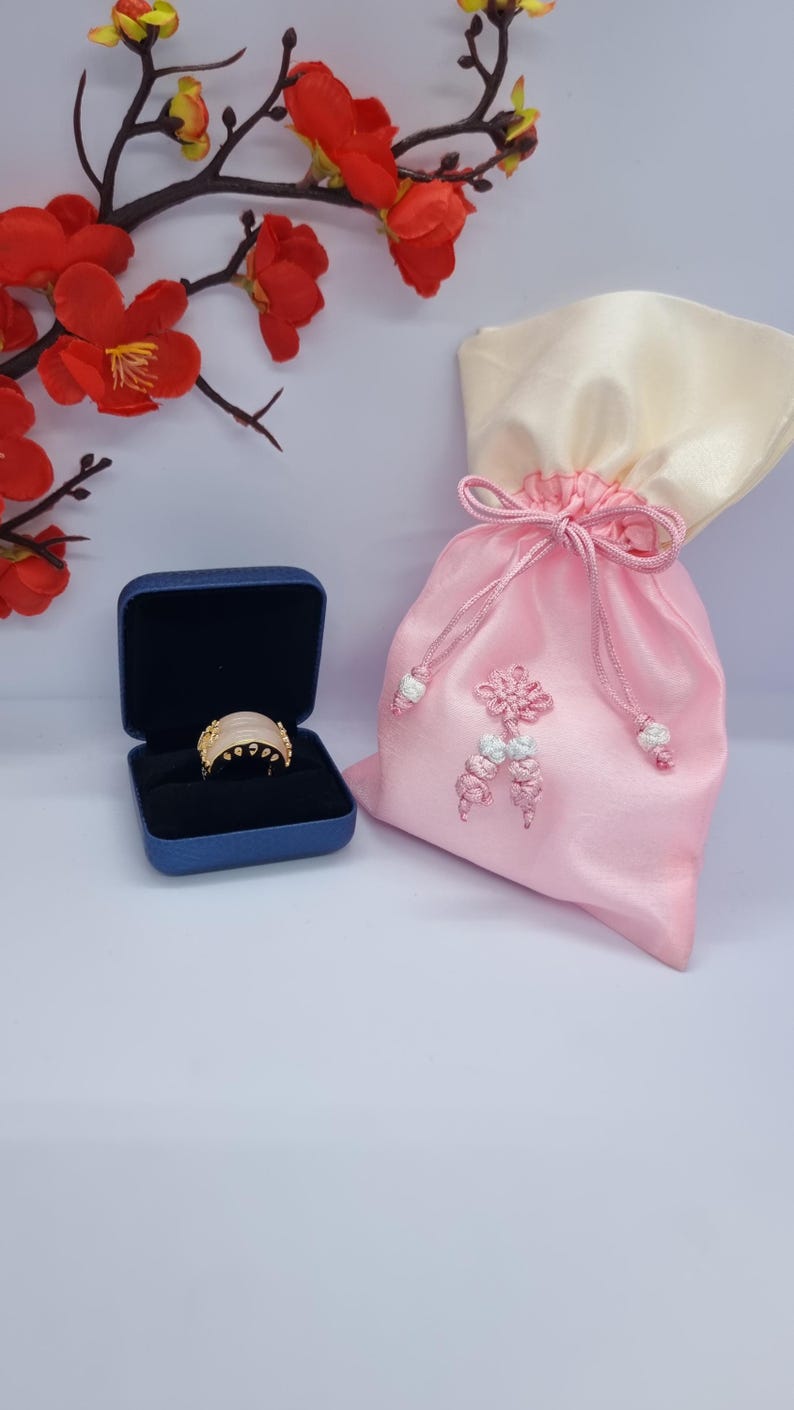 May include: A gold ring with a light pink stone is displayed in a blue ring box. A pink satin gift bag with a cream-colored top is next to the ring box. Red flowers and branches are in the background.