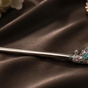 May include: A silver hair stick with a decorative end featuring a large, round, light green stone and floral accents in pink, blue, and white. The hair accessory rests on a dark brown, silky fabric.