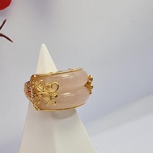 May include: A wide, rose-colored ring with gold filigree and accents. The ring has a rounded shape and is displayed on a white stand. The gold detailing includes floral and scroll designs, adding an ornate touch to the jewelry.