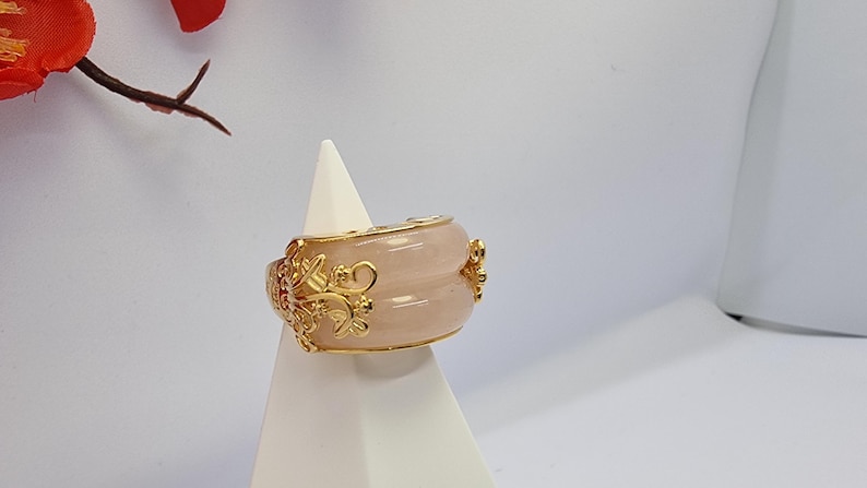 May include: A wide, rose-colored ring with gold filigree and accents. The ring has a rounded shape and is displayed on a white stand. The gold detailing includes floral and scroll designs, adding an ornate touch to the jewelry.