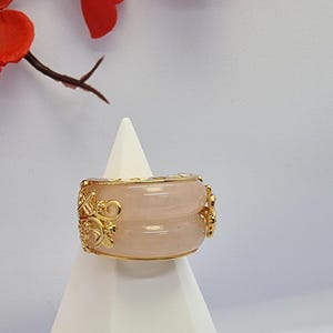 May include: A wide, light pink ring with gold filigree detailing. The ring is displayed on a white stand, with red flowers in the background. The ring has a double-band design.
