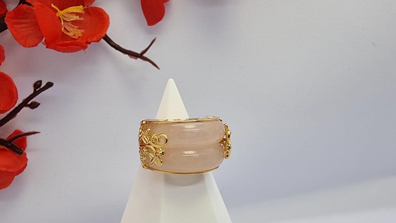 May include: A wide, light pink ring with gold filigree detailing. The ring is displayed on a white stand, with red flowers in the background. The ring has a double-band design.