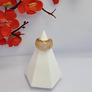 May include: A wide, light pink ring with gold-colored filigree detailing. The ring is displayed on a white geometric stand, with red flowers and branches in the background. The ring's design is symmetrical and ornate.