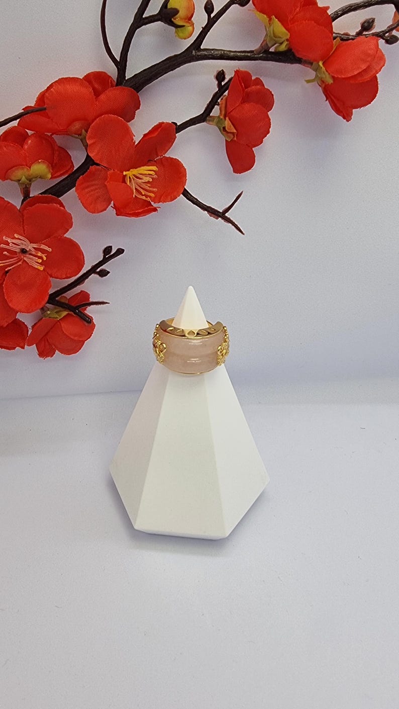 May include: A wide, light pink ring with gold-colored filigree detailing. The ring is displayed on a white geometric stand, with red flowers and branches in the background. The ring's design is symmetrical and ornate.