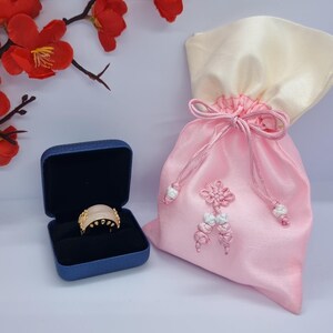 May include: A gold ring with a light pink stone is displayed in a blue jewelry box. A pink satin drawstring bag with a cream-colored top is next to the box. Red and yellow flowers are in the background.