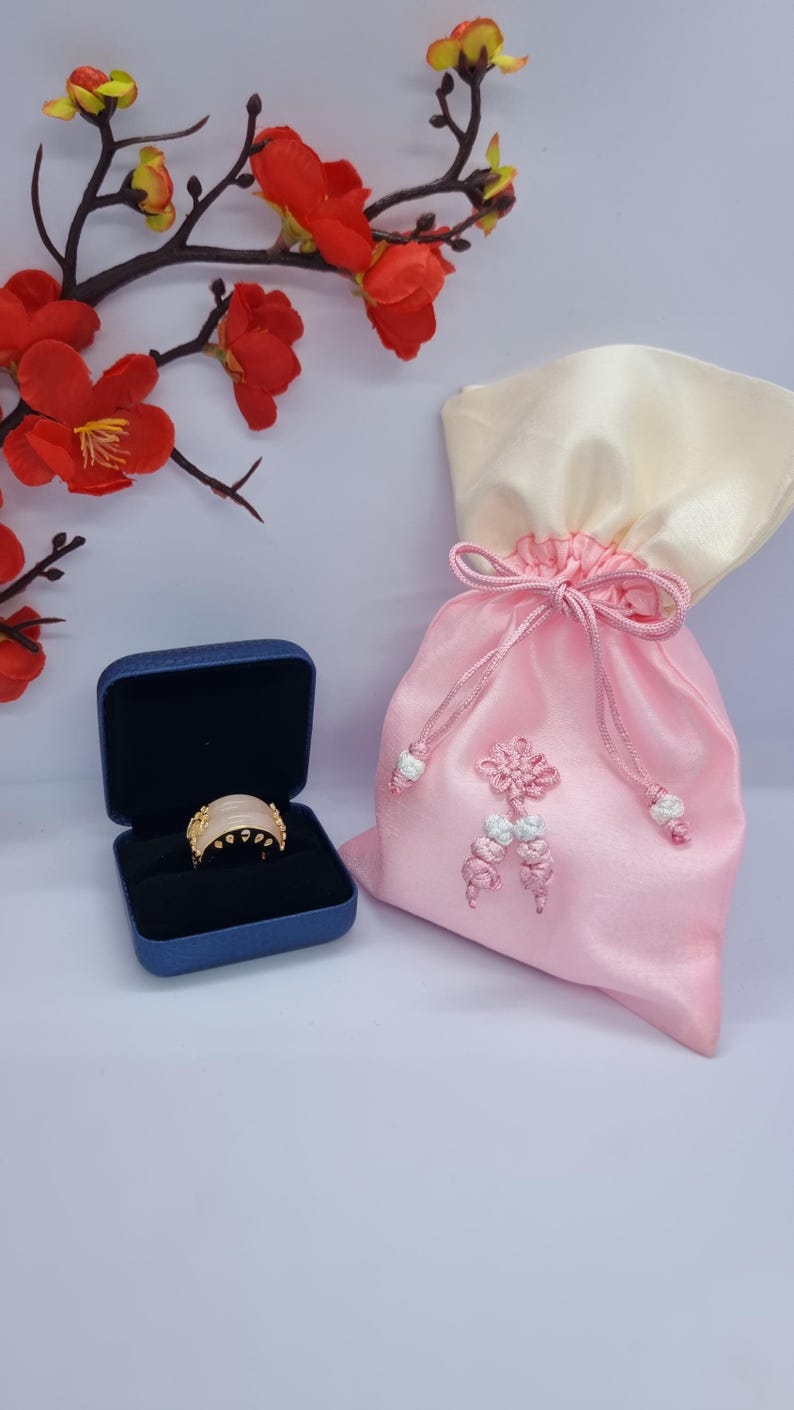 May include: A gold ring with a light pink stone is displayed in a blue jewelry box. A pink satin drawstring bag with a cream-colored top is next to the box. Red and yellow flowers are in the background.