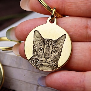 Può includere: Un portachiavi color oro con un ciondolo rotondo con un ritratto inciso di un gatto. Il gatto ha un motivo a strisce e tratti del viso dettagliati. Il portachiavi è tenuto da una persona.