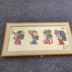 May include: A framed artwork featuring four colourful paper cutouts of women's heads with elaborate headdresses. The artwork is on a beige background with Chinese text in the bottom right corner.