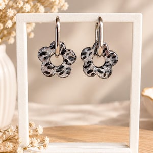 May include: A pair of silver-toned hoop earrings featuring flower-shaped charms. The charms have a black and silver leopard print pattern with a glittery finish. The earrings are suspended from a white rectangular display stand.