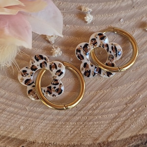 Puede incluir: Dos colgantes con forma de flor con un anillo dorado. Los colgantes tienen un diseño de estampado de leopardo con detalles de purpurina dorada. El fondo es una superficie de madera marrón claro con una flor rosa.