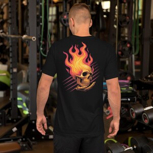 May include: Black short-sleeve t-shirt featuring a vibrant graphic of a skull engulfed in flames. The skull is depicted in shades of yellow and orange, with the flames extending upwards. The design is set against a backdrop of diagonal pink lines.