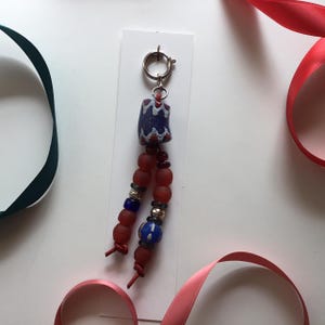 May include: A handmade beaded charm with a silver clasp. The charm features a dark blue and white patterned bead, red beads, and smaller blue and silver beads. Red ribbon is visible in the background.