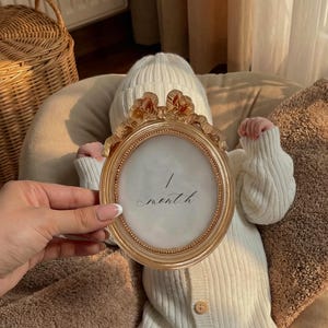 May include: A baby in a white knitted onesie and hat, resting on a brown cushion. A gold oval frame with the words "1 month" is held up. A wicker basket is visible in the background. The scene is bathed in soft light.