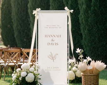 May include: A tall, rectangular white fabric wedding welcome sign suspended on a white metal frame with cream ribbons. The sign features the text "WELCOME TO THE WEDDING OF HANNAH & DAVIS", a floral line drawing, and the date "24.1.2026 KRYAL CASTLE".