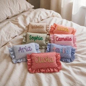 Puede incluir: Siete cojines rectangulares de cuadros vichy con bordes de volantes, cada uno con un nombre bordado diferente: Isabella, Lilith, Sophie, Camelia, Aria, Amelia y Olivia. Los cojines vienen en varios colores, incluyendo beige, naranja, verde, rosa, azul y rojo.