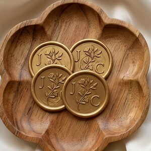 May include: A flower-shaped wooden dish containing four circular gold wax seals. Each seal features an embossed floral sprig design with the letters J and C flanking the stem. The wood grain is visible on the dish, which has six rounded petals.