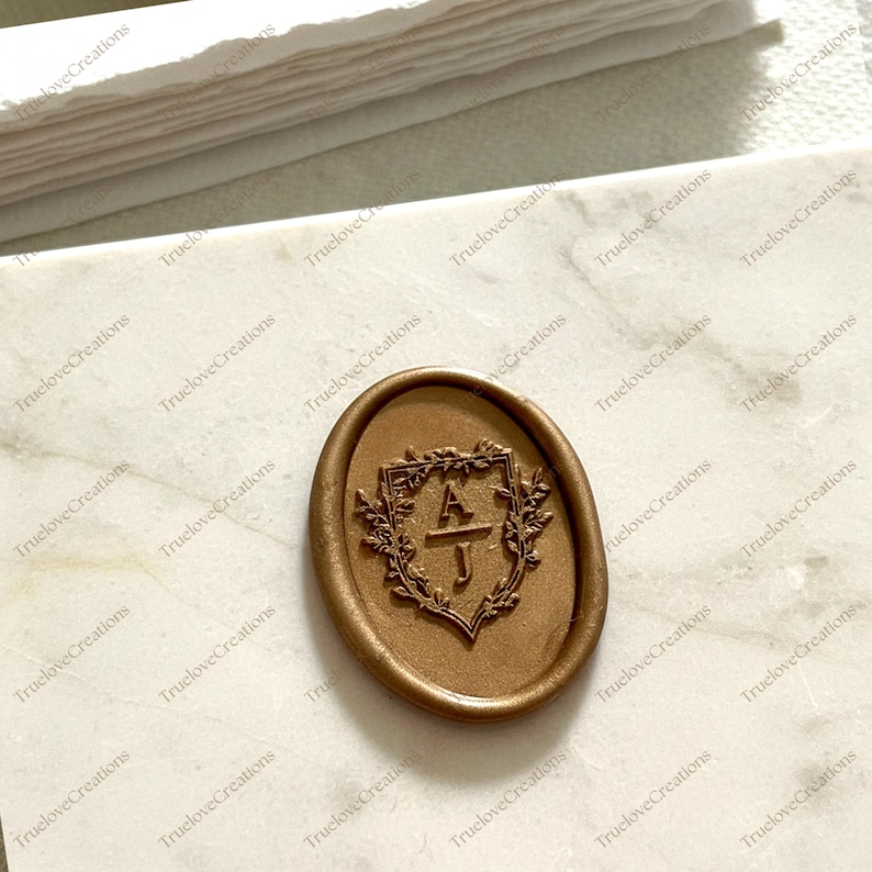 May include: A bronze-coloured wax seal featuring the initials "A J" within a decorative shield, framed by a floral wreath. The oval seal rests on a marble surface, with the text "TrueloveCreations" repeated in the background.