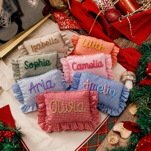 May include: A collection of seven small rectangular fabric pillows with ruffled edges and a gingham pattern in various colours. Each pillow features a name embroidered in cursive script: Isabella, Lilith, Sophie, Camelia, Aria, Amelia, and Olivia.