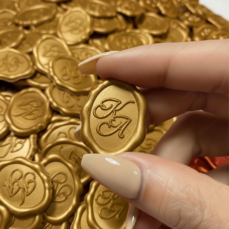 May include: Gold wax seals, each featuring an embossed KA monogram within an oval frame. The seals have a raised edge and a polished surface. One seal is held up, with a pile of similar seals in the background. These are suitable for adding a refined touch to stationery or craft projects.
